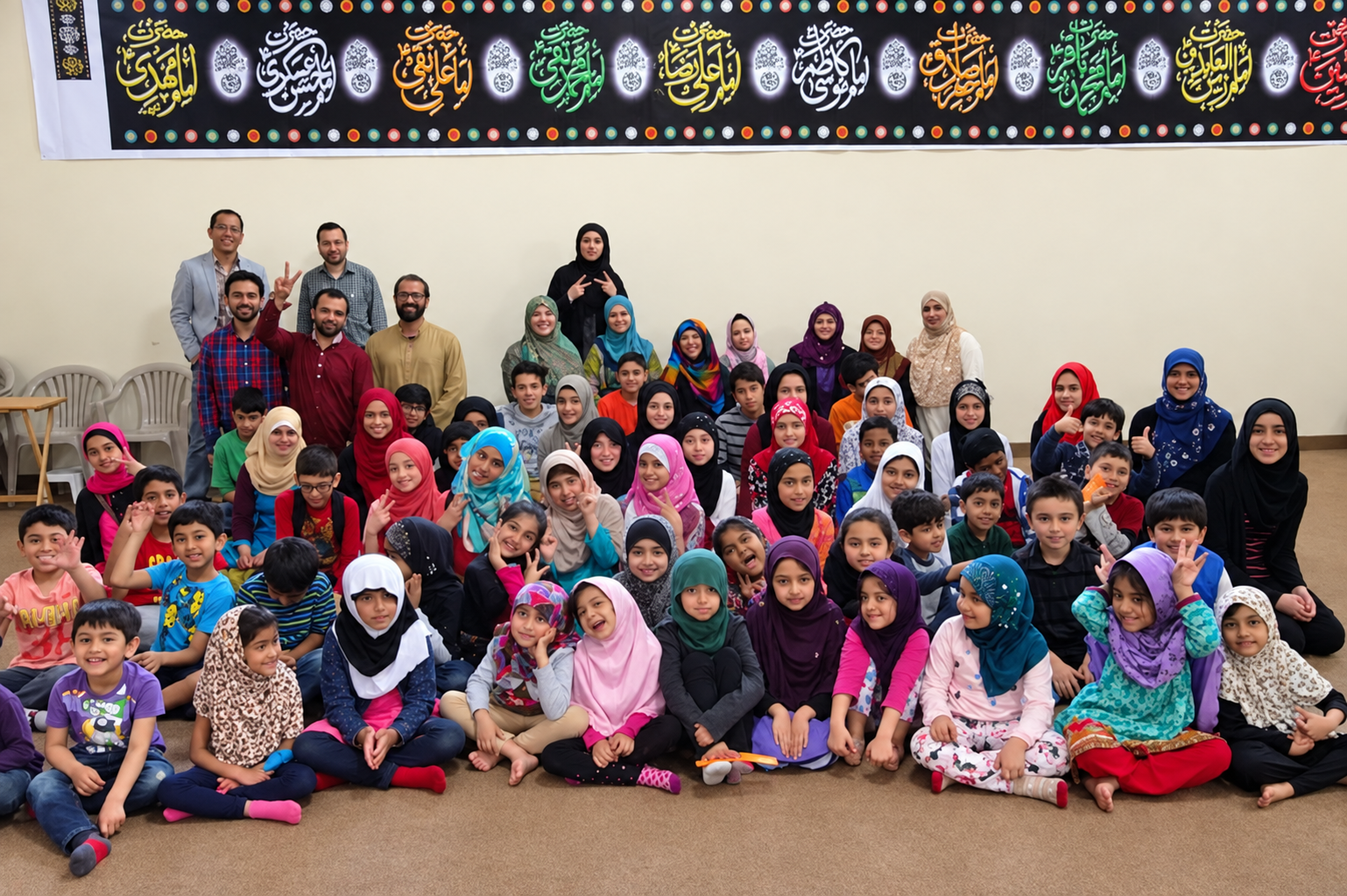 Large group photo of students and teachers beneath the names of the Ahlul Bayt calligraphy banner